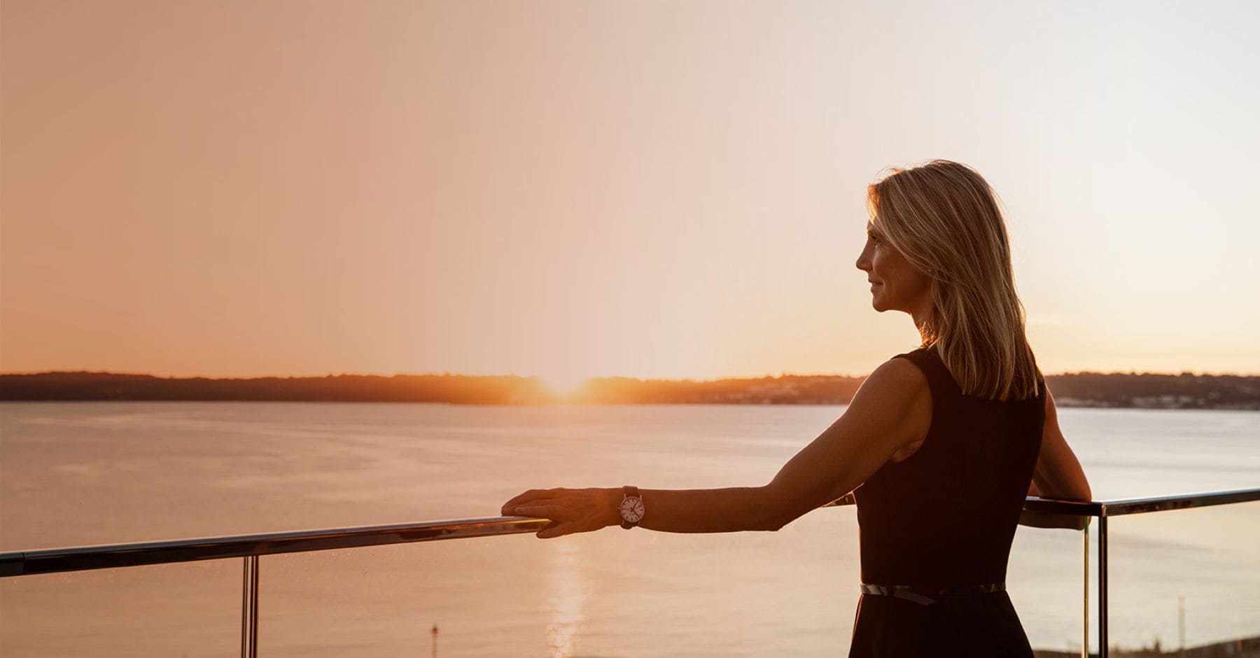 Businesswoman standing on office balcony overlooking St Aubin's bay at sunset